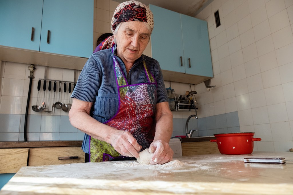 An elderly woman in the kitchen is kneading dough on a floured wooden surface.