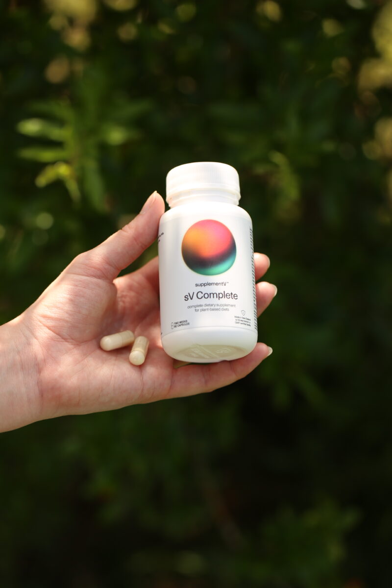 The photo shows a person's hand holding up a bottle of supplementV's sV Complete and two capsules. sV Complete is an all-in-one vegan supplement that the company claims is more than just a multivitamin.